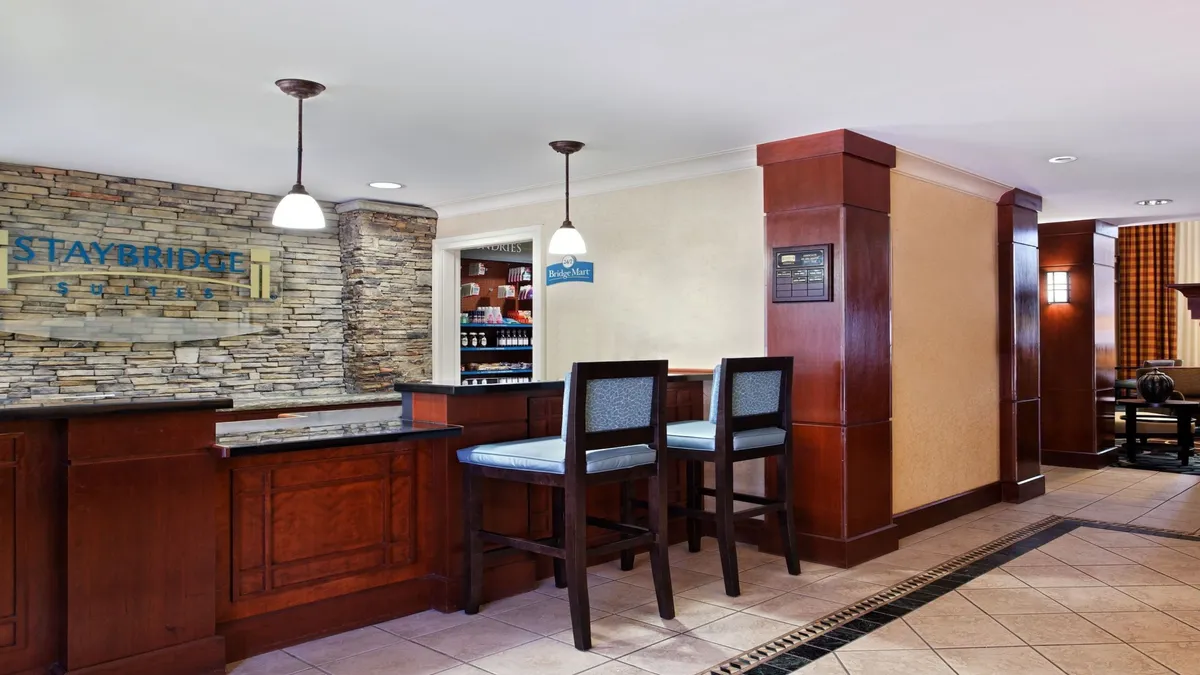 A warm and inviting reception area with a stone wall, hanging lights, bar stools, and a modern reception desk. The space is abuzz with guests checking in, chatting casually with the friendly staff, and briefly relaxing as they arrive or depart.