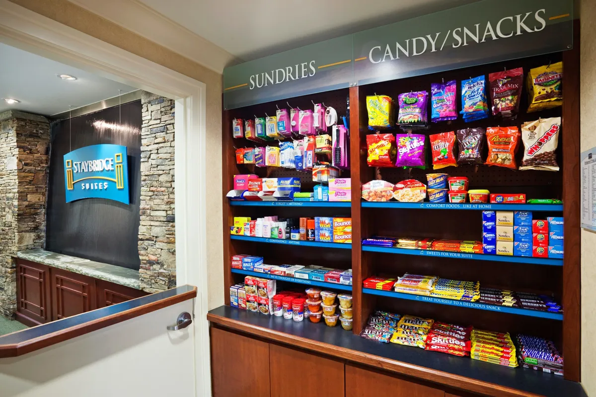 A cozy snack area featuring shelves stocked with a variety of treats, a welcoming Staybridge sign, and a stone accent wall. The layout encourages guests to stop by for a quick bite or beverage and socialize in a comfortable, informal setting.