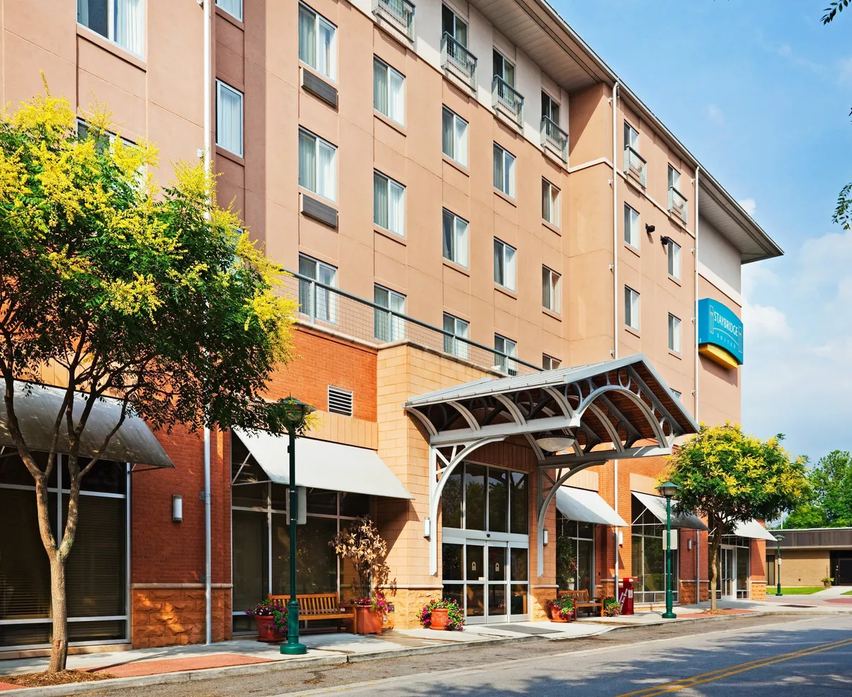 This exterior shot showcases the inviting entrance awning, attractive landscaping, and an outdoor seating area where guests can be seen relaxing and interacting. In the foreground, a group of guests is checking in, while others enjoy the outdoor space, reflecting the accessibility of nearby restaurants and social venues.