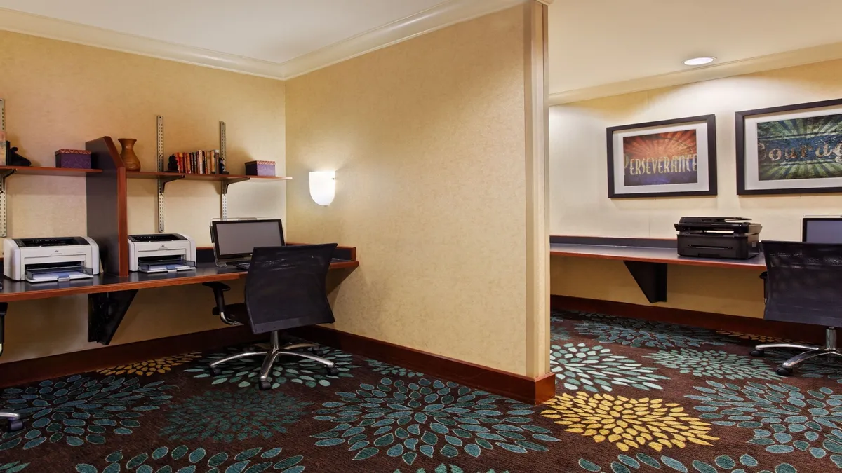 A modern business center with multiple computer desks, printer stations, colorful wall art, and decorative carpet. Guests are seen working, printing documents, and collaborating in a focused, productive environment.