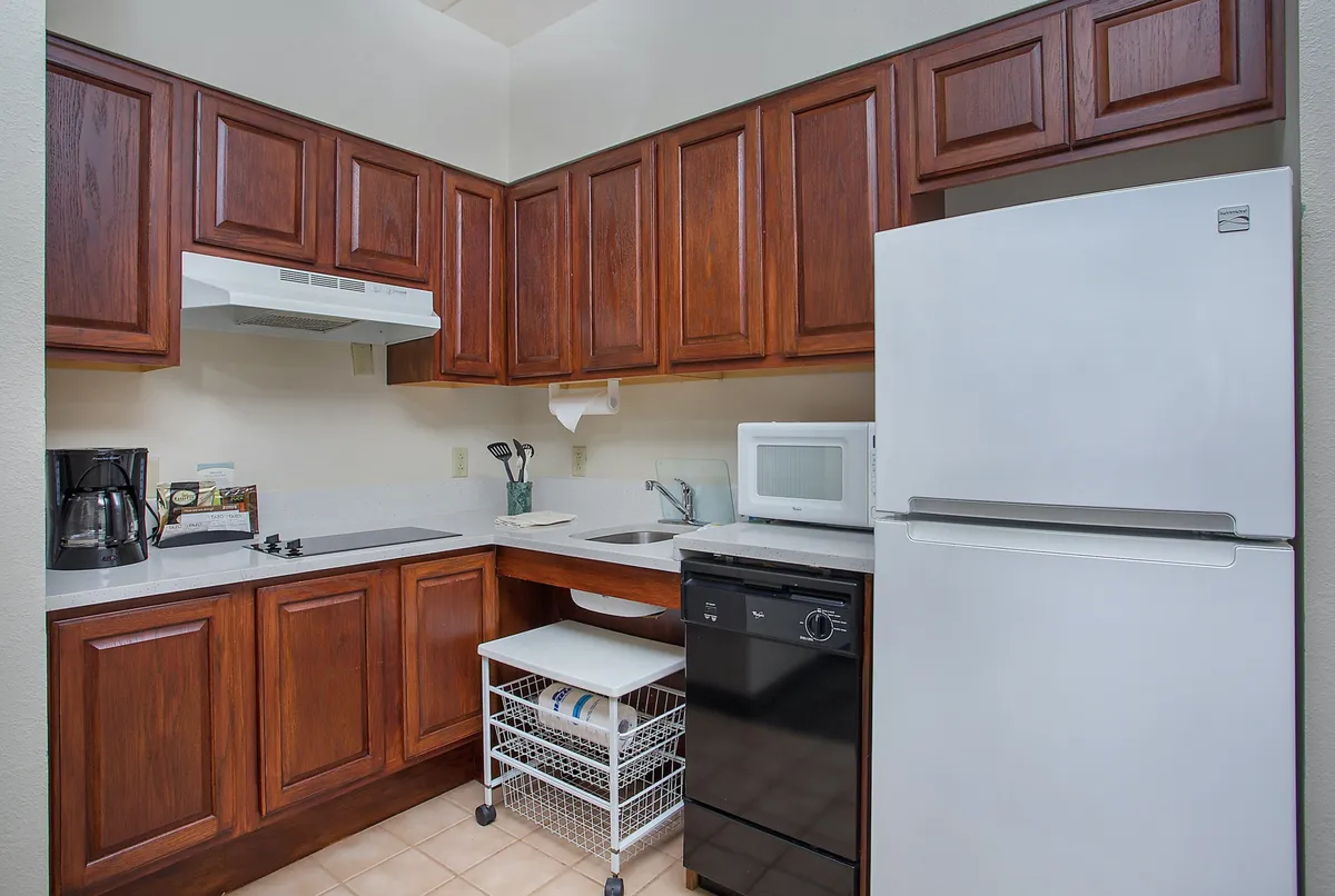 A warmly lit kitchen featuring wooden cabinets, white appliances including a stove and microwave, a spacious sink area, and a practical kitchen cart. The layout suggests plenty of room for meal prep, with social touches indicating gatherings or shared meals.